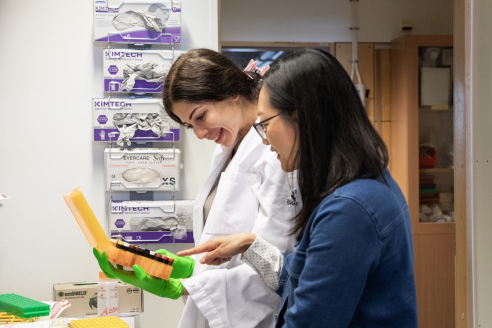 Två kvinnor i labbrockar samarbetar, ler och tittar på en färgglad låda.