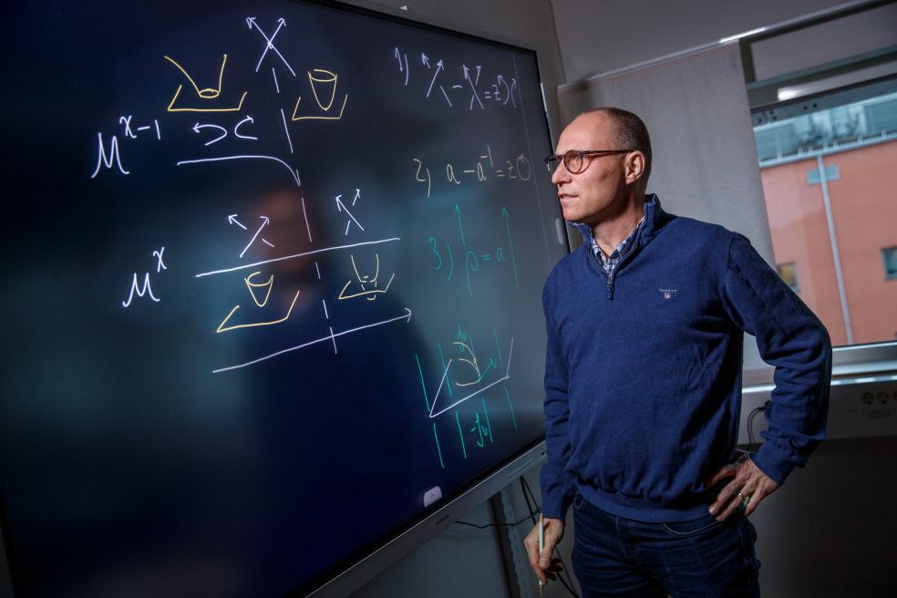 En man i glasögon står vid en stor skärm med matematiska formler.