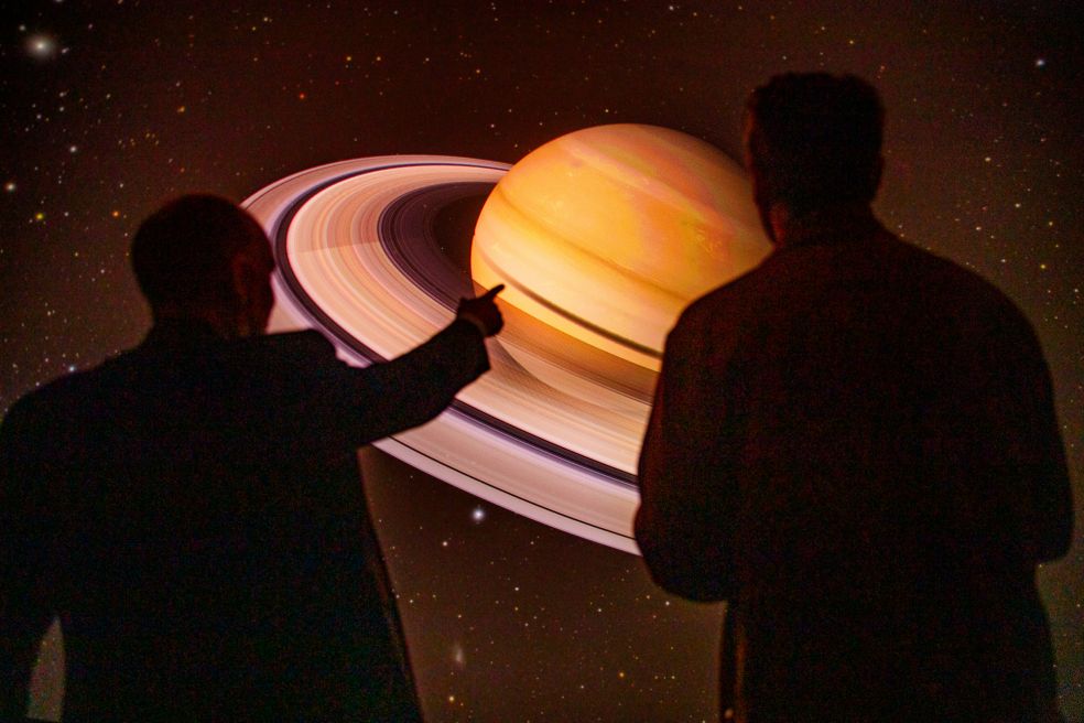 Två personer står och pekar på en stor bild av planeten Saturnus.
