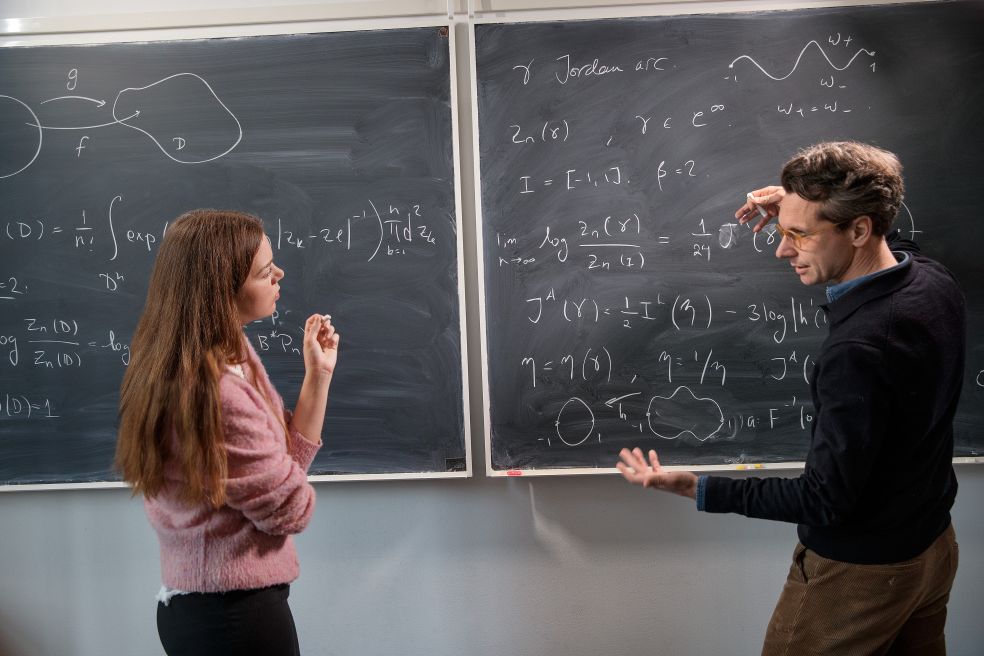 Kvinna och man diskuterar matematik framför svarta tavlor med ekvationer.