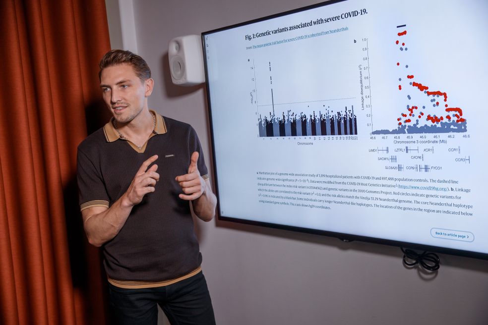 En man står framför en TV-skärm som visar diagram och text, gestikulerar.