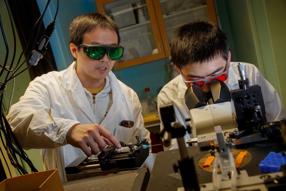 Två forskare i skyddsglasögon arbetar med en mikroskop på ett laboratorium.
