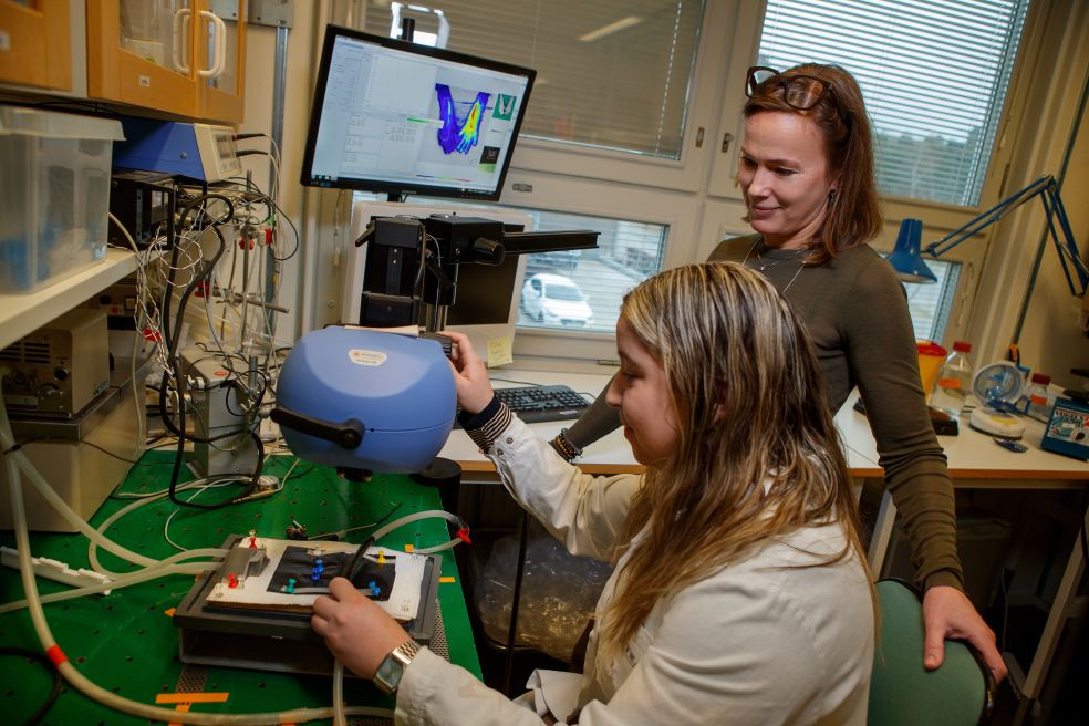 Två kvinnor i labbutrustning, en arbetar vid en apparat och en observerar.