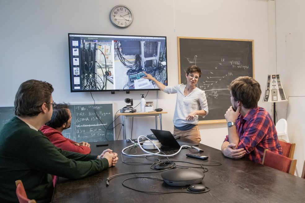 En person presenterar information för tre åhörare i ett rum med skolplanscher.