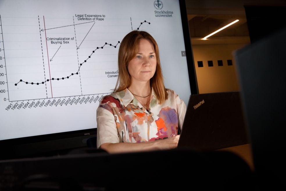 Kvinna med lång hår i färgglad skjorta framför en graf på en skärm.