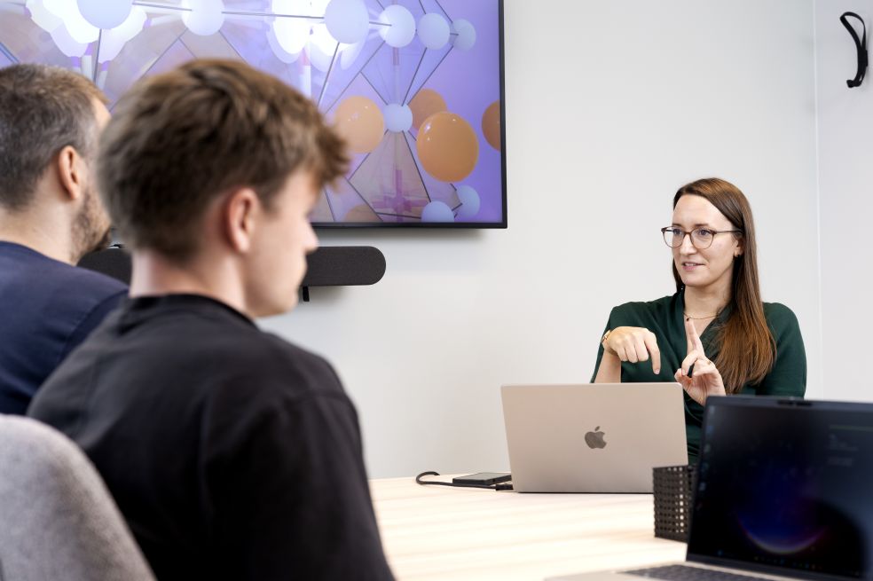 Kvinna presenterar vid ett mötesbord med två män som lyssnar.