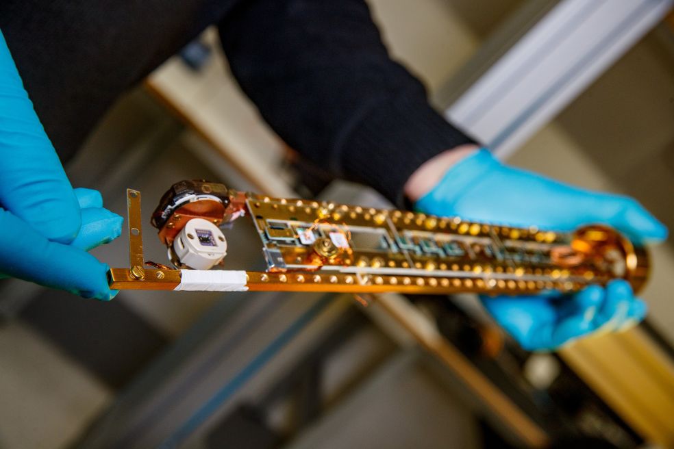 En person med blå handskar håller en lång, teknisk komponent med kretskort.