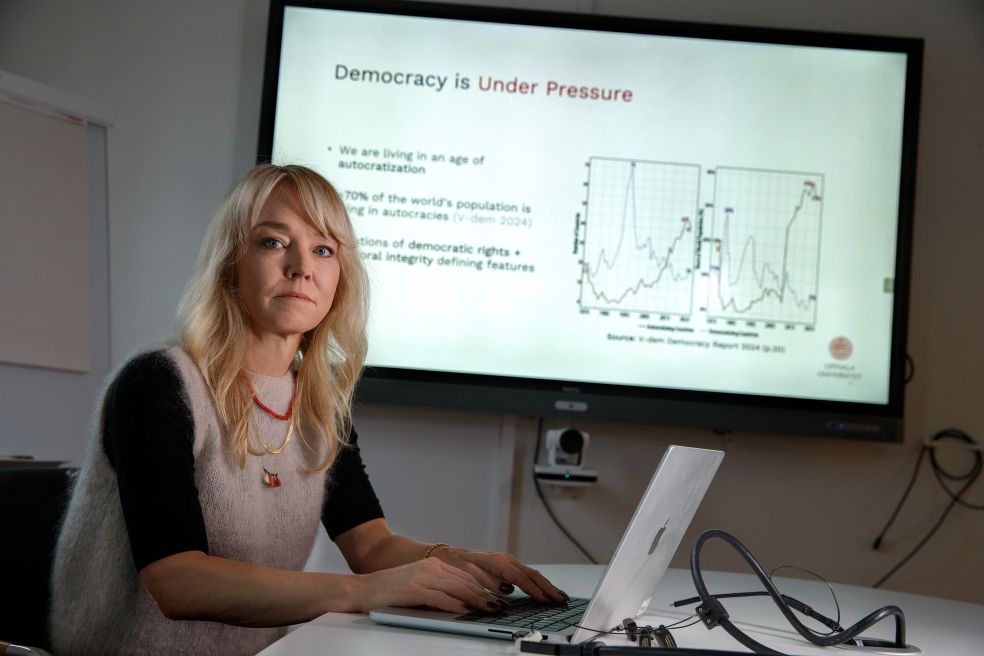 Kvinna med blont hår sitter vid datorn framför en stor skärm som visar grafer.