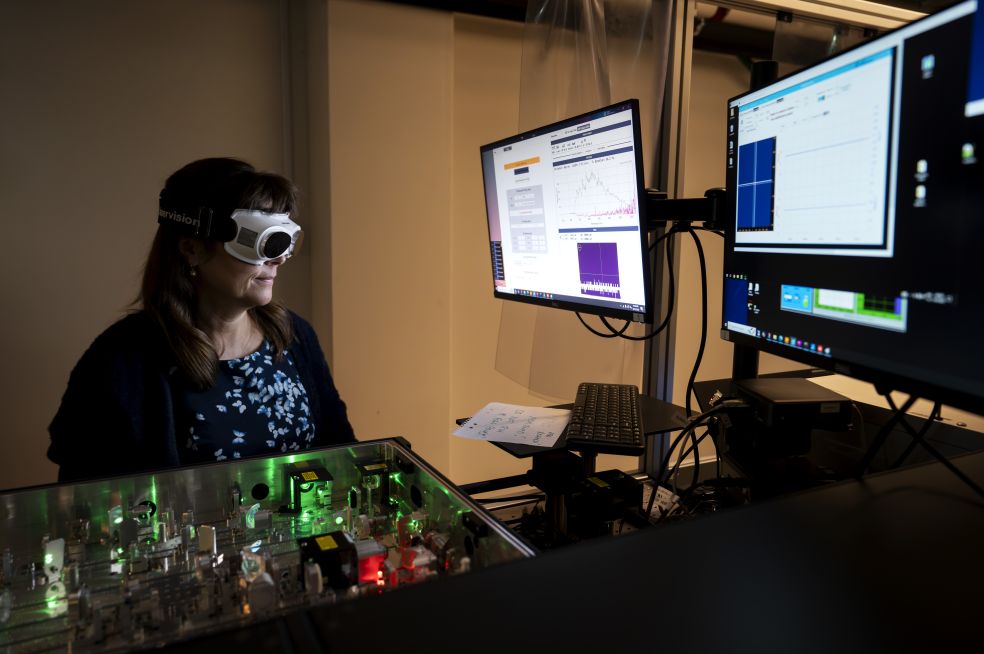 Kvinna med skyddsglasögon arbetar vid dator med flera skärmar och labbutrustning.