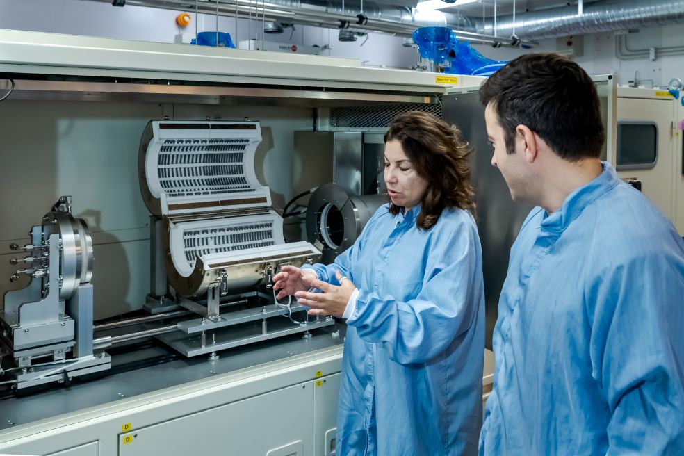 Två personer i labbrockar framför en avancerad maskin, diskuterar dess funktion.