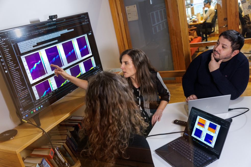 Tre personer diskuterar grafik på en stor skärm i ett kontorsutrymme.