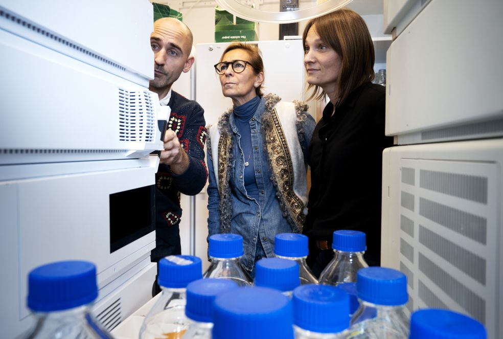 Tre personer tittar på en kyl i en butik, flaskor med blåa lock syns i förgrunden.