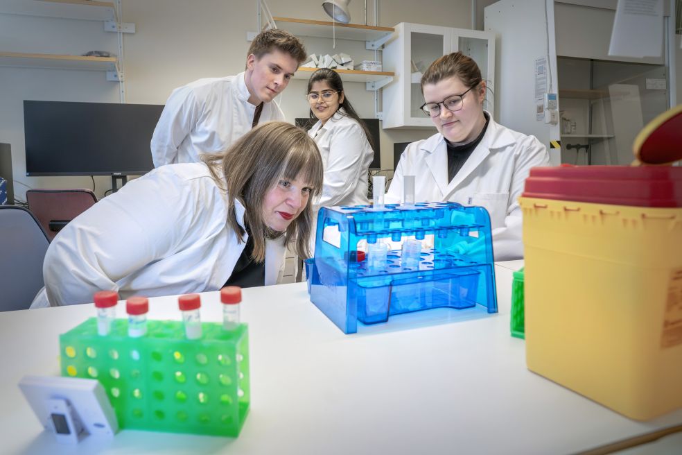 Fyra personer i labbrockar inspekterar ett experiment i ett laboratoriemiljö.