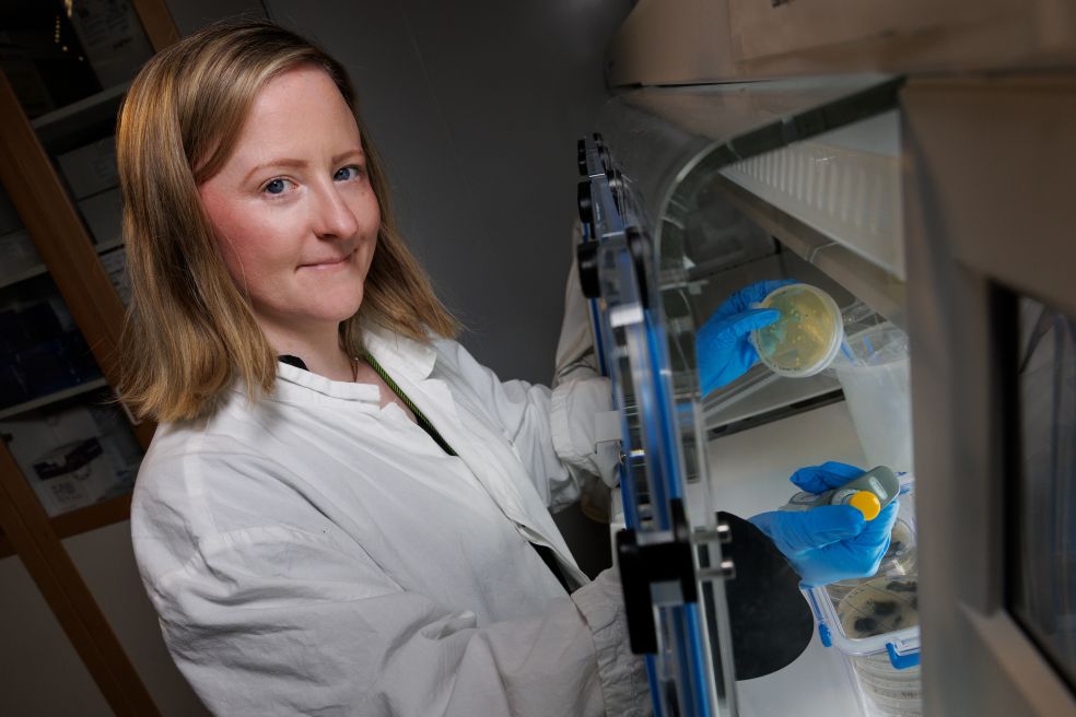 Kvinna i laboratorierock arbetar vid en bänk med prover i en kontrollerad miljö.