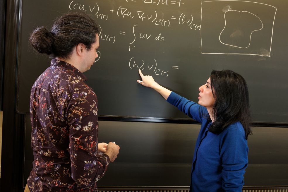 Två personer diskuterar matematik vid en tavla med formler och en skiss.