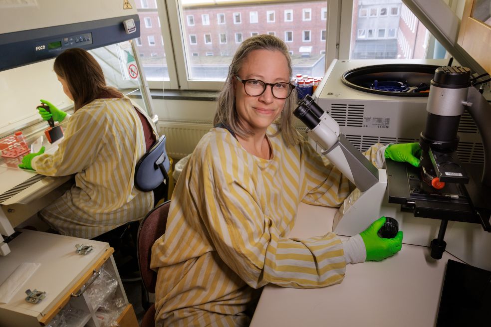 Kvinna med glasögon och grön handskar arbetar vid mikroskop i laboratorium.