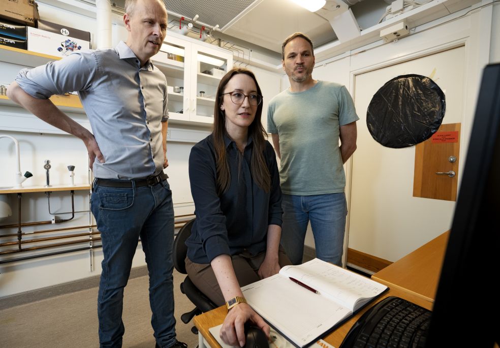 Tre personer står och sitter vid en dator i ett rum med arbetsmaterial.