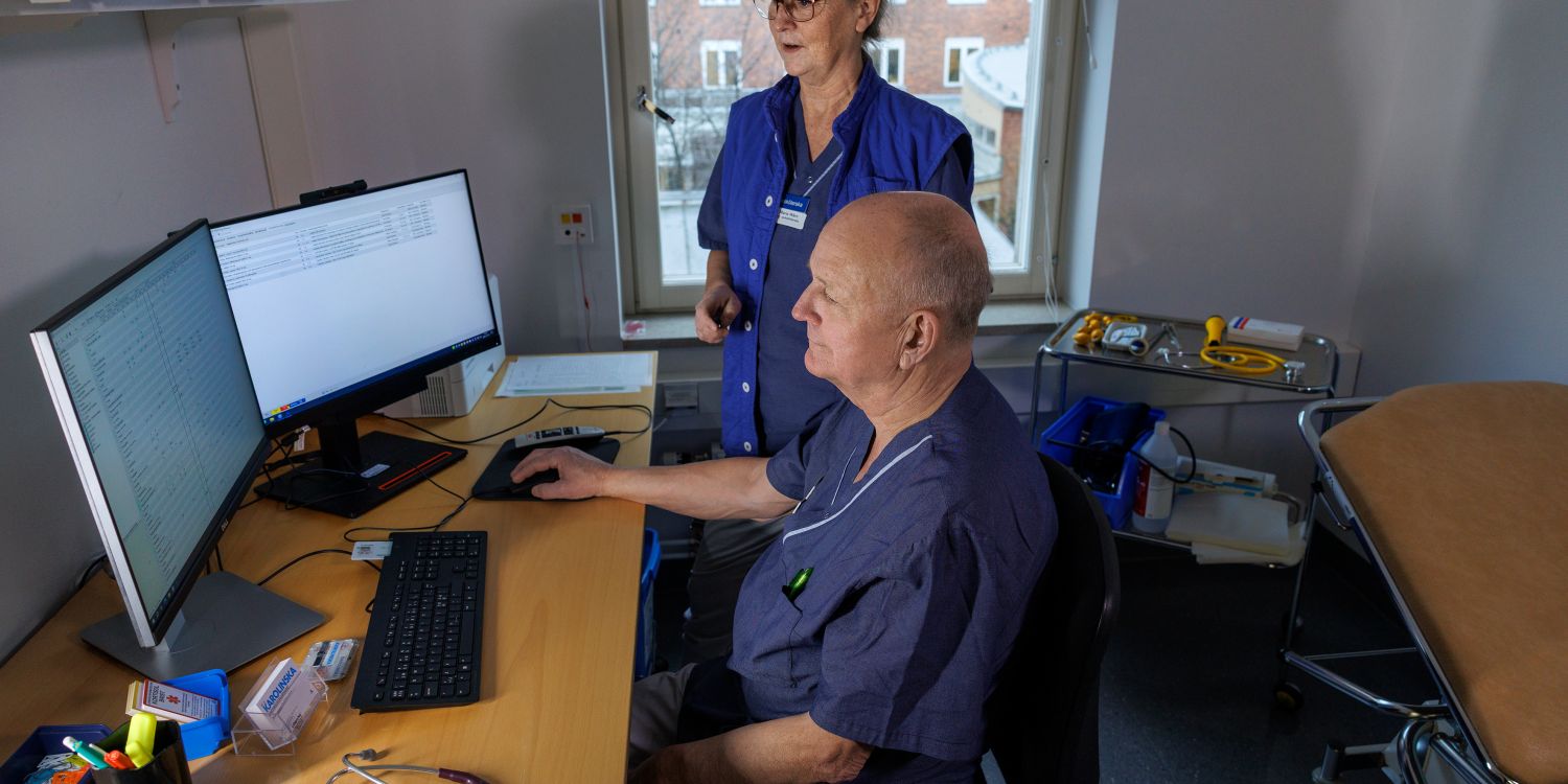 Sjuksköterska står bakom en patient vid dator i ett vårdrum.