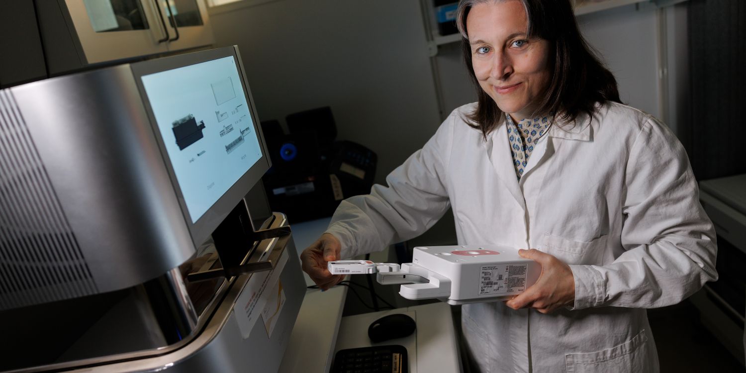 Kvinna i labbrock håller en apparat framför en dator i ett laboratorium.