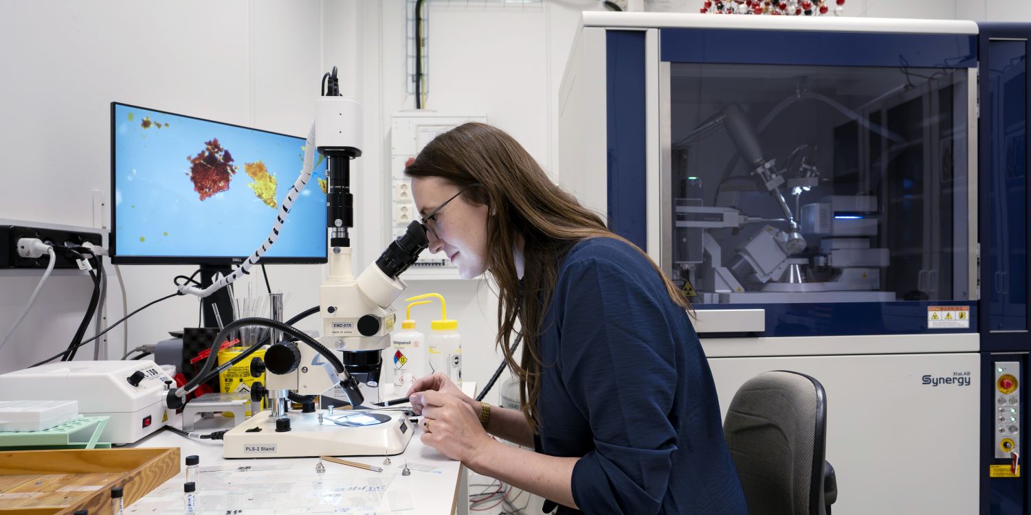 Kvinna som arbetar vid mikroskop i ett laboratorium med en datorskärm i bakgrunden.
