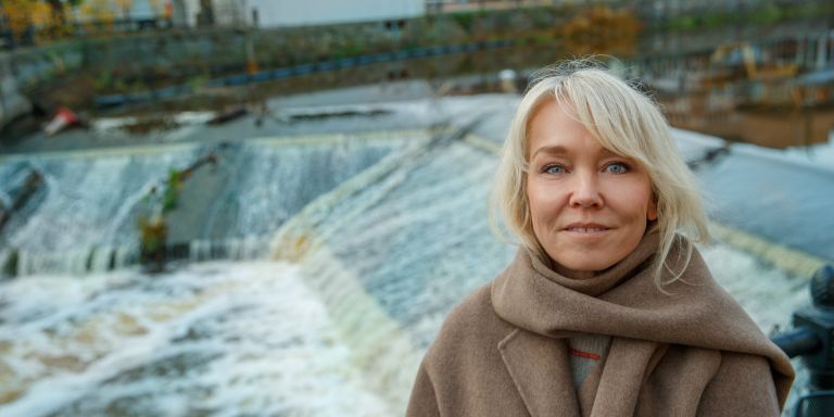 Kvinna med blont hår i en brun kappa, står framför en forsande vattenström.