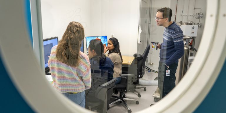 Fyra personer i ett labb en står utanför och tittar in genom ett fönster.