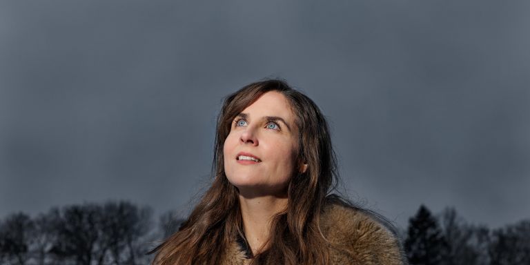 Kvinna med lång brunt hår och pälsjacka, blickar upp mot en mörk himmel.