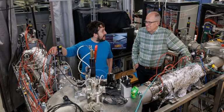 Två män diskuterar vid ett laboratorium med avancerad utrustning och rörsystem.