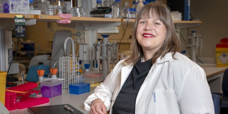 Kvinna med labbrock ler framför en laboratoriumsarbetsbänk med utrustning.