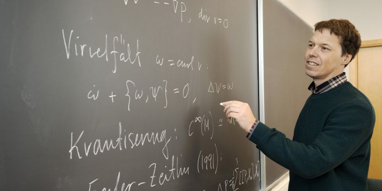 En lektor pekar på matematiska formler på en svart tavla.
