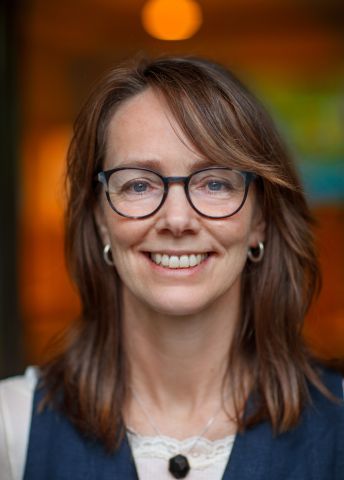 Leende kvinna med glasögon och brunt hår, i en mjukt upplyst miljö.