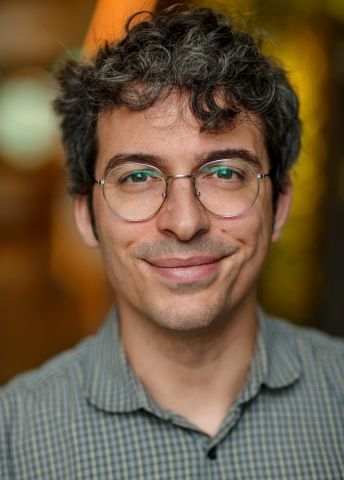 En leende man med glasögon och lockigt hår, i en mjukt upplyst miljö.