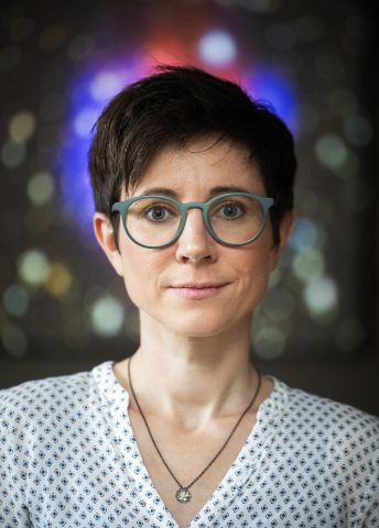 Kvinnovisa med kort hår, glasögon och vit prickig skjorta, med färgglad bakgrund.