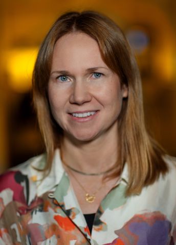 Kvinna med långt, brunt hår klädd i färgglad skjorta, ler mot kameran.