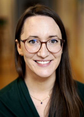 Leende kvinna med glasögon, långbrunt hår och grön tröja, fotograferad inomhus.