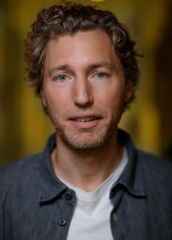 En man med lockigt hår och en grå skjorta mot en suddig bakgrund.