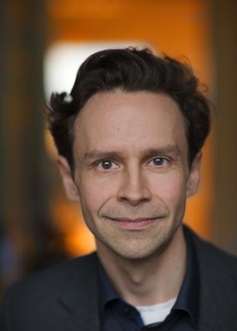 Man med ljus hy och mörkt hår, ler framför en suddig, varm bakgrund.