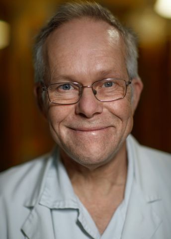 Leende man med glasögon, iförd vit labbrock, mot en varm bakgrund.