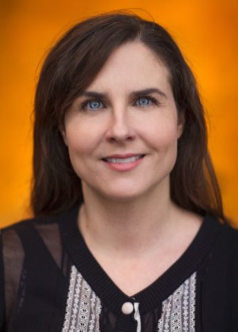 Kvinna med mörkt hår och blå ögon, klädd i en svart tröja mot en orange bakgrund.