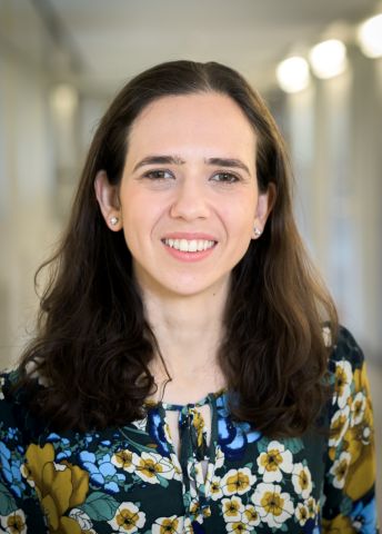 Kvinna med långt, brunt hår och blommig blus, ler i en ljus korridor.