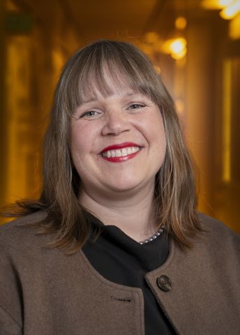 Leende kvinna med långt hår i en brun kappa, i en varm inomhusmiljö.