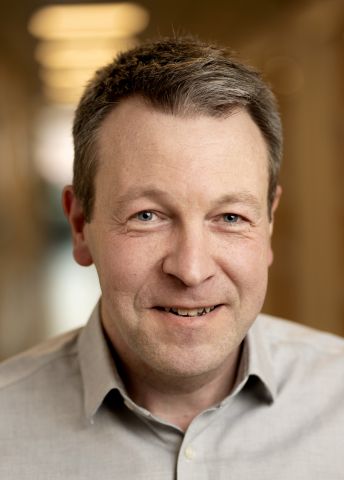 Leende man med grått hår, iförd skjorta, stående i en ljus korridor.