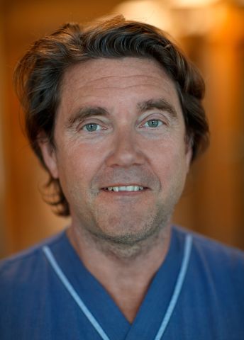 Man med långt brunt hår i en blå tröja, leende mot kameran. 