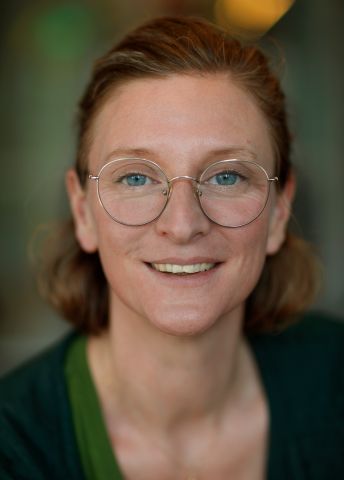 Leende kvinna med glasögon, kort hår och grön tröja i suddig bakgrund.