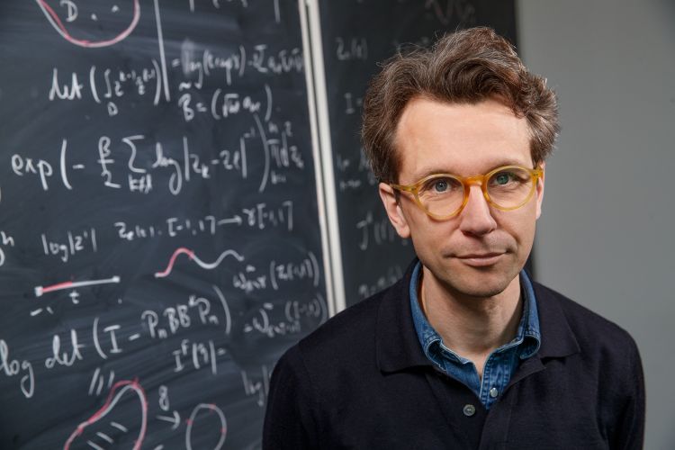 En man med glasögon står framför en svarts tavla med matematiska formler.