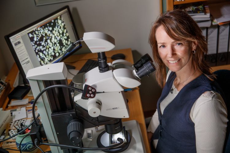 Kvinna ler bredvid ett mikroskop, med en dator i bakgrunden.