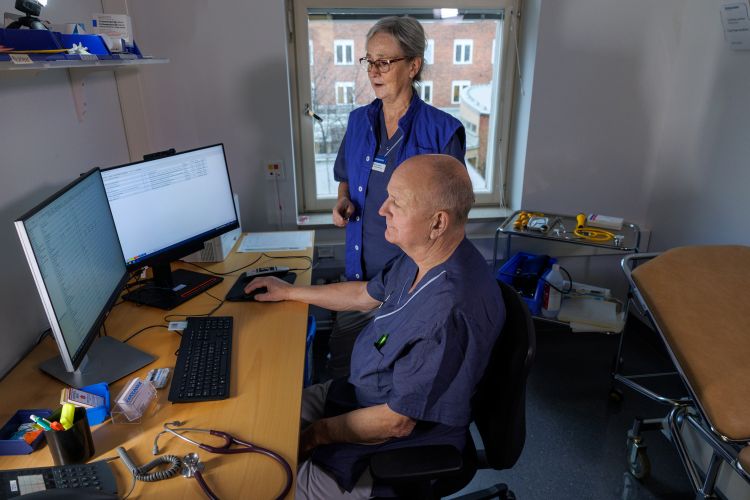 Sjuksköterska står bakom en patient vid dator i ett vårdrum.