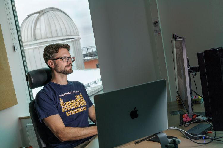 Man sitter vid skrivbordet framför en dator med observatorium i bakgrunden.