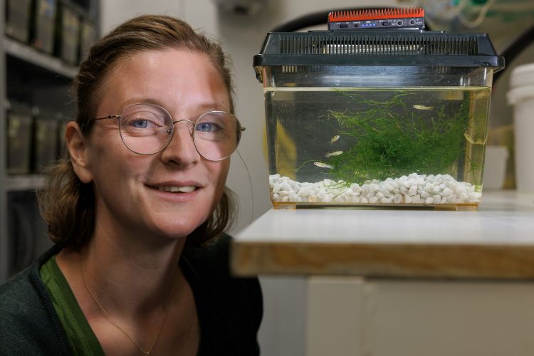 Leende kvinna med glasögon sitter vid ett akvarium med gröna växter och fiskar.