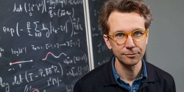 En man med glasögon står framför en svarts tavla med matematiska formler.